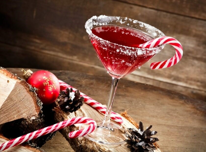 Christmas cocktails concept image, a picture of a martini glass with a red drink inside, sugar around the rim and a candy cane hanging off the side. Lying around the cocktail is wooden logs, pinecones, a red bauble and candy canes. 