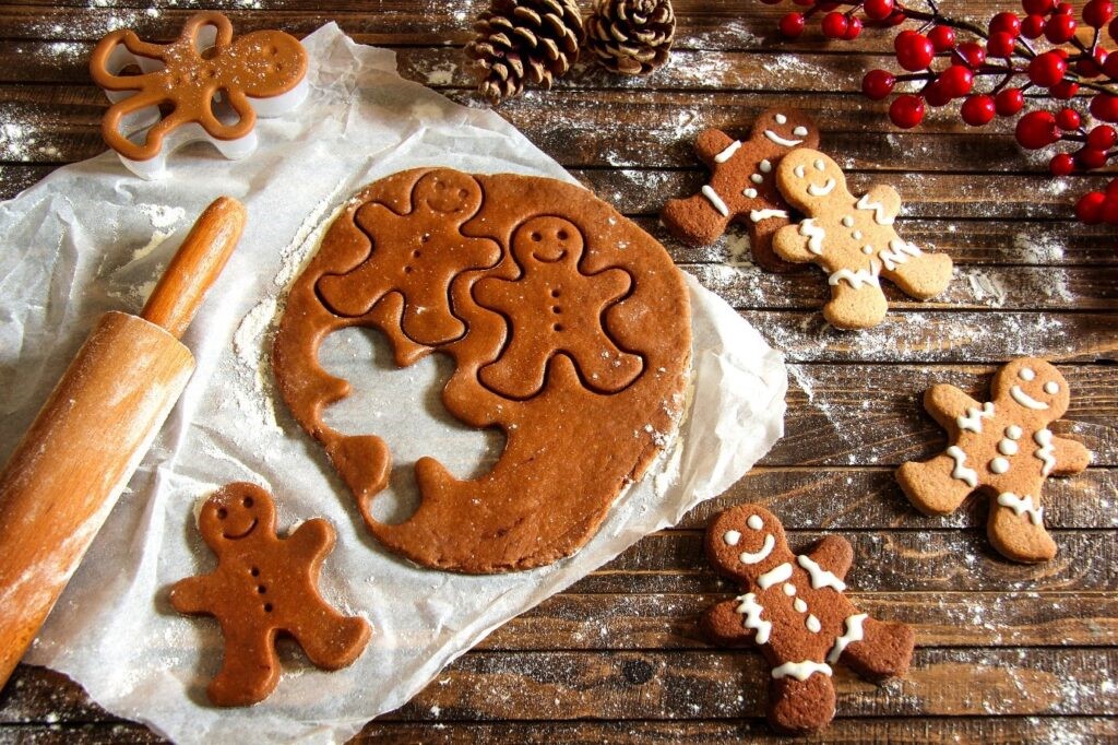 Image of gingerbread men being made, the gingerbread dough is rolled out next to a rolling pin and gingerbread men are being cut out using a cutter. The dough is surrounded by iced gingerbread men.