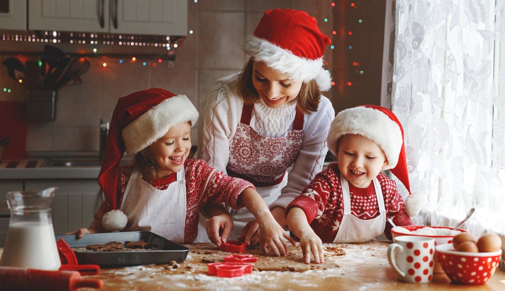 Image of a mother and her two children baking biscuits in the kitchen at Christmas time. They're wearing Christmas jumpers, Christmas aprons and Santa hats.