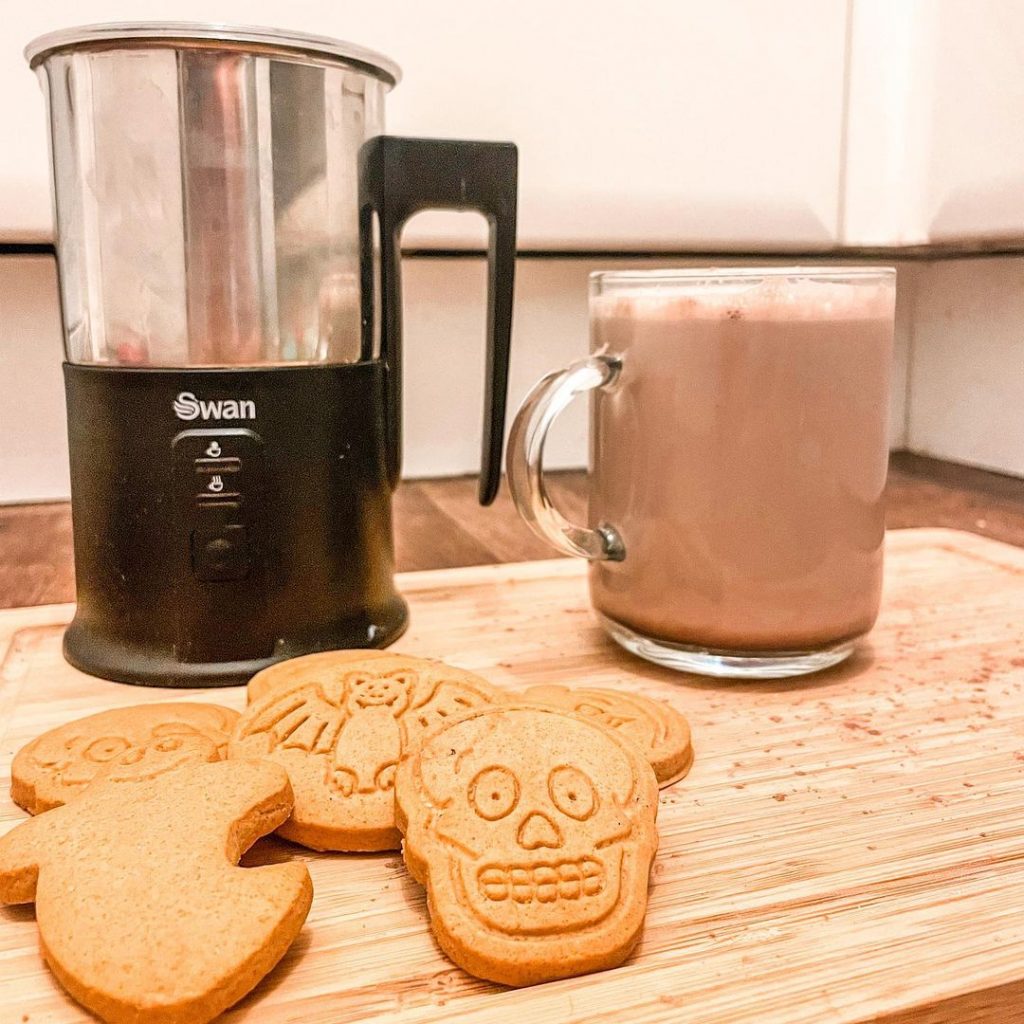 Swan Automatic Milk Frother next to a small hot chocolate and homemade halloween biscuits on a wooden chopping board