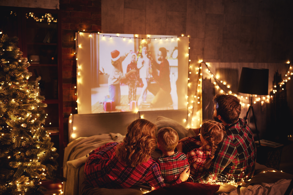 Family spend christmas day watching films on projector screen with fairy lights all around them