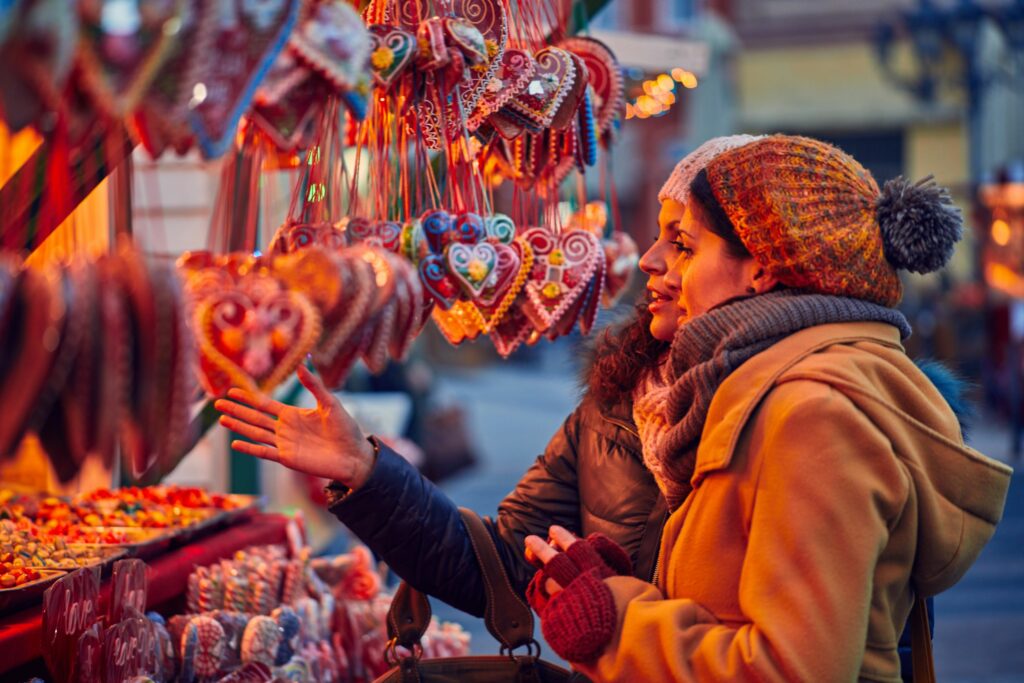 A colour image of two women in hats, scarves and gloves at a Christmas market. They're in front of a market stall selling Christmas decorations on blog about festive things to do. 