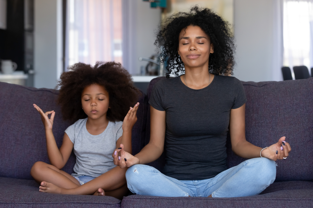 Mother and daughter sit side-by-side, cross legged on a couch in a meditation pose with eyes closed for blog about healthy lifestyle changes.
