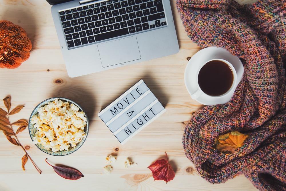 Birdseye view of a laptop, blanket and cup of coffee next to a side of popcorn and a lightbox that reads "movie night"